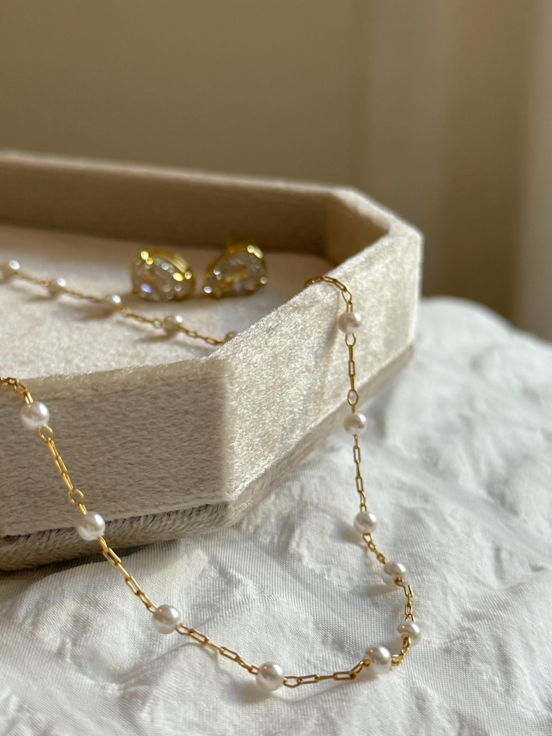 May include: A gold chain necklace featuring small white pearls spaced evenly along the links, accompanied by a pair of gold stud earrings with clear, faceted stones. The jewellery pieces are displayed together in a light beige velvet tray.