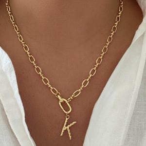 May include: Gold-colored chain necklace with a pendant featuring the letter "K". The necklace has a textured, braided design on the letter and a clasp closure. The chain is made of oval links.