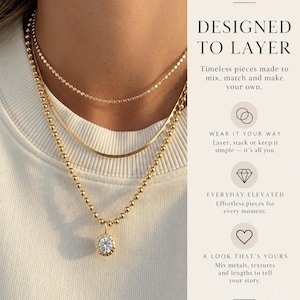 May include: Three gold-toned necklaces layered together. The top is a delicate chain, the middle is a flat snake chain, and the bottom is a beaded chain with a round pendant featuring a clear stone. Text reads: DYLAN RAE DESIGNED TO LAYER.