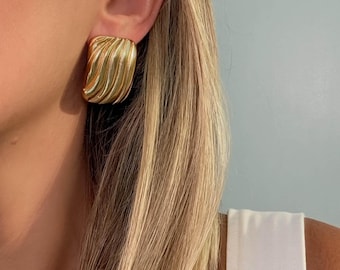 May include: A gold-toned earring featuring a rectangular shape with a series of vertical, rounded ridges. The surface has a polished, reflective finish that catches the light, creating a textured appearance on the earlobe.