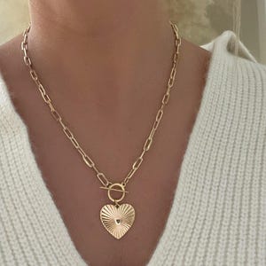 K&ouml;nnte beinhalten: Eine goldfarbene Halskette mit einem herzf&ouml;rmigen Anh&auml;nger. Der Anh&auml;nger hat ein strukturiertes Sonnenstrahl-Design und ein kleines Loch unten. Die Halskette hat einen Knebelverschluss und eine Kette mit rechteckigen Gliedern.
