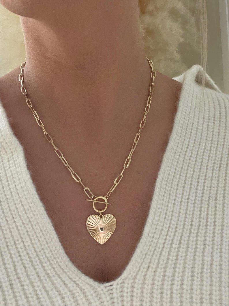 K&ouml;nnte beinhalten: Eine goldfarbene Halskette mit einem herzf&ouml;rmigen Anh&auml;nger. Der Anh&auml;nger hat ein strukturiertes Sonnenstrahl-Design und ein kleines Loch unten. Die Halskette hat einen Knebelverschluss und eine Kette mit rechteckigen Gliedern.