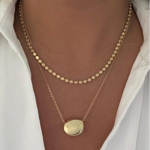 May include: Two-strand gold necklace. The top strand features small, round, flat gold beads. The bottom strand has a gold pendant with a smooth, rounded, irregular shape. The necklace is worn over a white collared shirt.