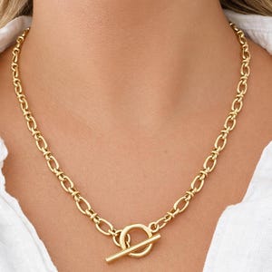 May include: A gold-colored chain necklace with an oval link design. The necklace features a toggle clasp closure. The necklace is worn against a white shirt.