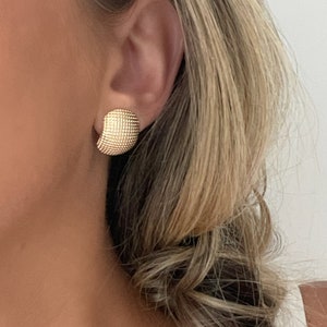 May include: Gold-colored stud earring with a textured, grid-like surface. The earring is a rounded square shape and is worn on the ear. The image shows the earring in close-up, highlighting its design and color. The earring is part of a jewelry collection.