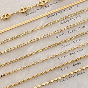 May include: A collection of eight gold-toned metal chains labeled Dainty Puff Mariner, Dainty Herringbone, Dainty Figaro, Dainty Paper Clip, Dainty Curb, Dainty Box Link, Dainty Rope, and Dainty Flat Bead, arranged in parallel rows on a light surface.