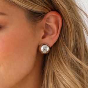 May include: Close-up of a silver, spherical stud earring. The earring is highly polished and reflects the surrounding environment. The earring is a simple, yet elegant accessory.