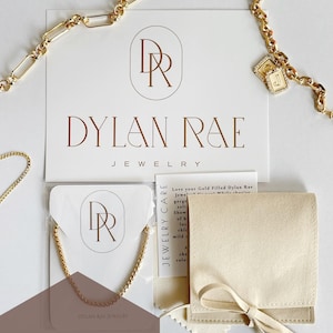 May include: Gold-filled chain necklace with a small square charm with the letter "J" on it. The necklace is packaged on a white card with the Dylan Rae Jewelry logo and a white microfiber pouch with a care card. The text "Love your Gold Filled Dylan Rae Jewelry" is visible on the care card.