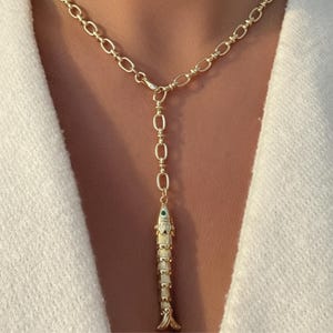 May include: A gold-toned lariat necklace featuring a textured chain and a vertical fish-shaped pendant. The fish pendant is detailed with a small, circular green accent near the head and a segmented body design, hanging from a central link.