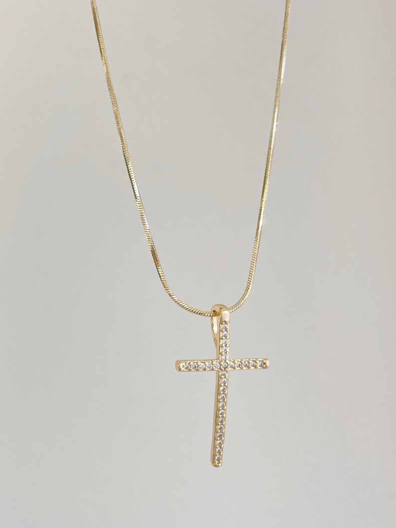 Puede incluir: Un collar de cadena dorada con un peque&ntilde;o colgante de cruz brillante. La cruz est&aacute; engastada con piedras de circonita c&uacute;bica transparentes.