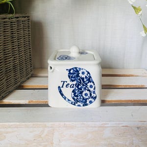 May include: A white ceramic tea container with a lid, featuring a blue floral cat design and the word "Tea". The container is rectangular with rounded edges and sits on a white wooden surface. Decorative flowers and a wicker basket are in the background.