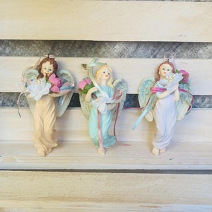 May include: Three porcelain angel figurines with wings, each holding a flower bouquet. The angels are wearing pastel blue, pink, and white dresses with ribbons.