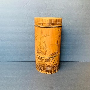 May include: A cylindrical bamboo vase with a carved design of two fish swimming amongst reeds. The vase is a light brown color and has a natural, rustic look.