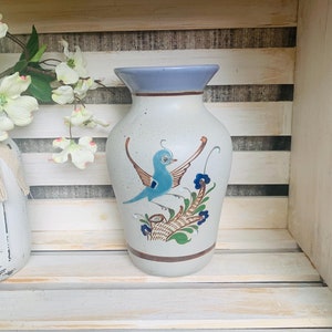 May include: A white ceramic vase with a blue rim and a painted design of a blue bird with green and brown foliage. The vase is sitting on a wooden shelf.