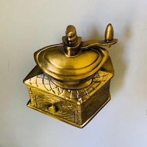 May include: A gold-coloured, vintage-style coffee grinder. It has a square base with a drawer, a central grinding mechanism, and a hand crank. The design incorporates wood-grain texture and decorative elements.