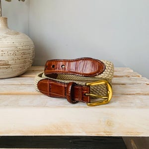 Puede incluir: Un cintur&oacute;n trenzado beige con detalles de cuero marr&oacute;n y una hebilla dorada. El cintur&oacute;n presenta un dise&ntilde;o texturizado y un estilo cl&aacute;sico. El cuero tiene un patr&oacute;n de cocodrilo.