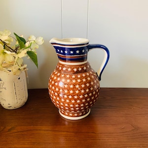 May include: A ceramic pitcher with a patriotic design. The top features a blue band with white stars and red stripes, while the body is brown with white stars. It has a blue handle and a white interior. A decorative piece for the kitchen or dining room.