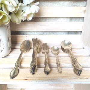 May include: A collection of vintage silverware, including spoons and forks, arranged on a light-colored wooden surface. The silverware has ornate detailing and a silver finish. A vase of white and pink flowers is in the background.