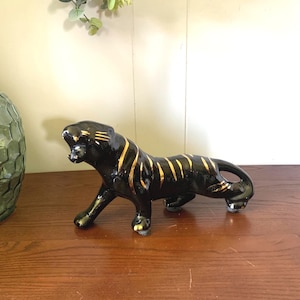 May include: A glossy black ceramic tiger figurine featuring gold painted stripes along its body. The tiger is depicted in a prowling stance with its head turned slightly to the side, showcasing a smooth, reflective finish and detailed gold accents.