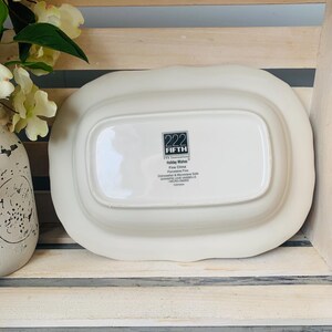 May include: White oval serving dish with a raised edge and a scalloped rim. The bottom of the dish has a black label with the text "222 FIFTH" and "Holiday Wishes Fine China Dishwasher & Microwave Safe Garantie Lave-Vaisselle MICRO-ONDES"