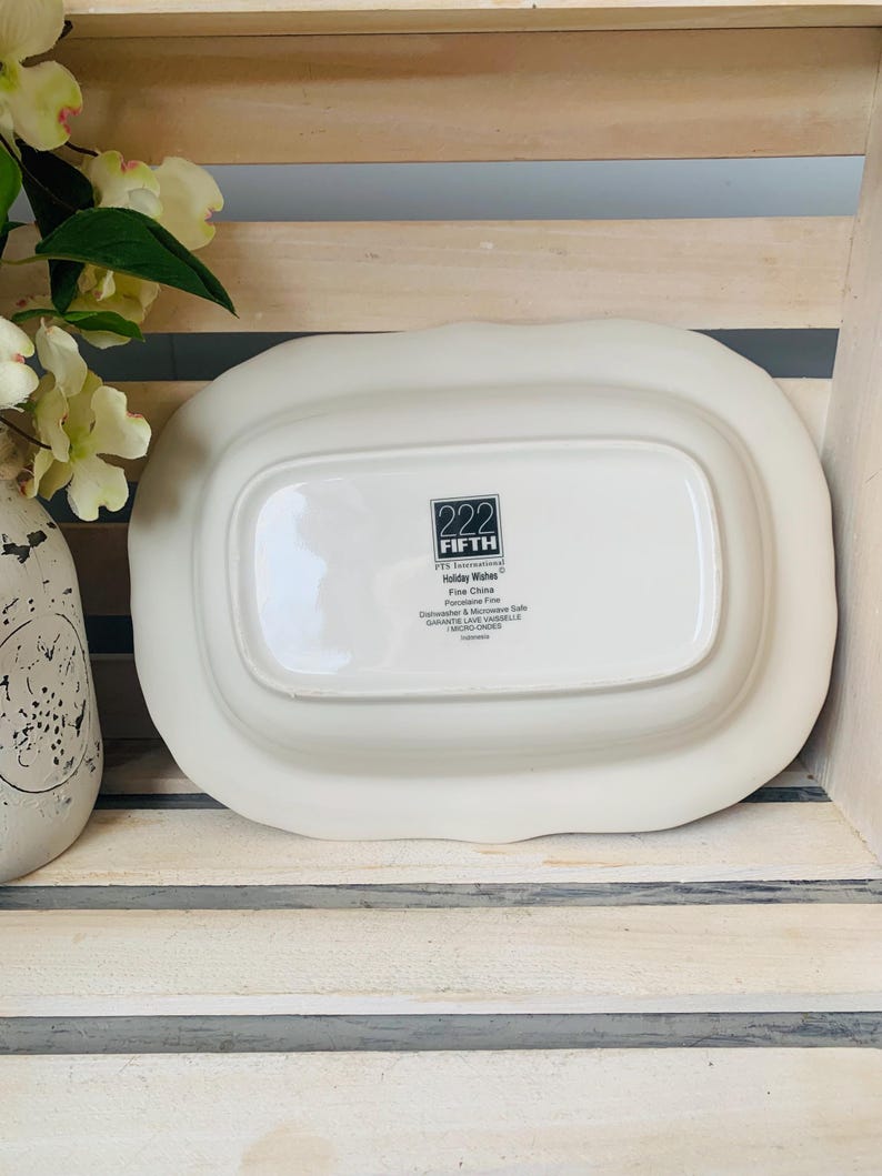 May include: White oval serving dish with a raised edge and a scalloped rim. The bottom of the dish has a black label with the text "222 FIFTH" and "Holiday Wishes Fine China Dishwasher & Microwave Safe Garantie Lave-Vaisselle MICRO-ONDES"