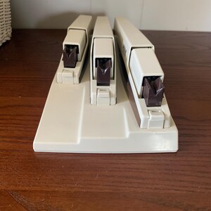 May include: A set of three off-white staplers with dark brown stapling mechanisms. The staplers are arranged on a matching base, with the largest stapler on the right. The set is designed for office or home use.