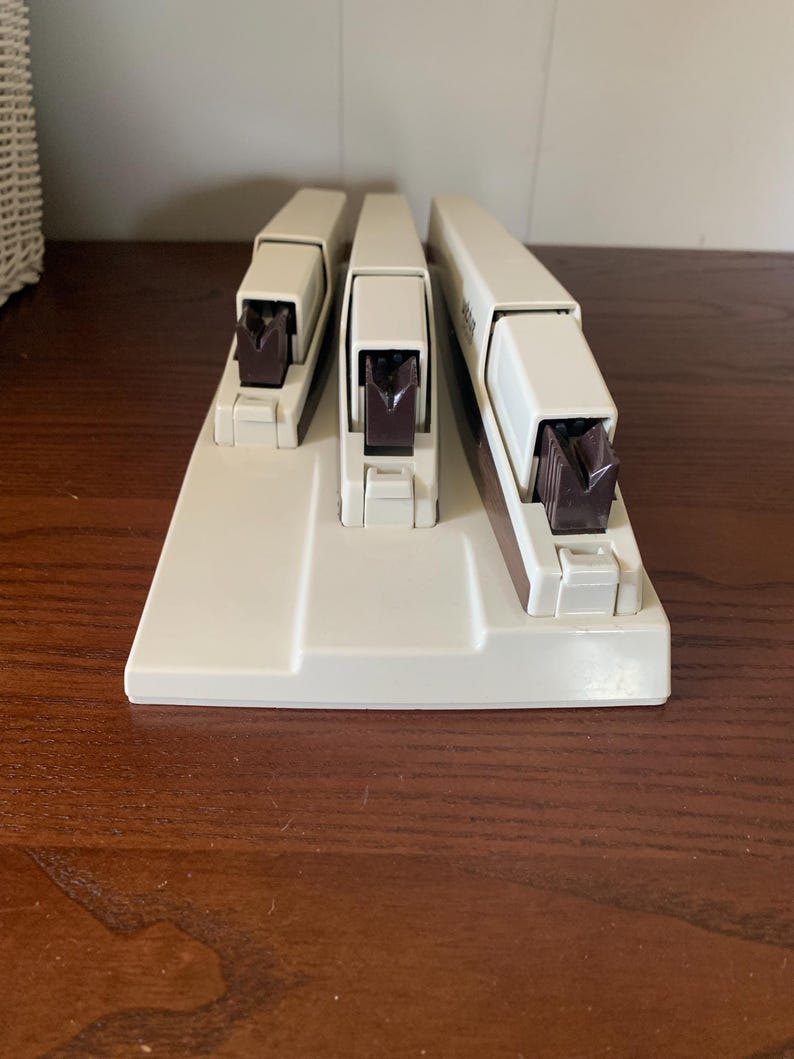 May include: A set of three off-white staplers with dark brown stapling mechanisms. The staplers are arranged on a matching base, with the largest stapler on the right. The set is designed for office or home use.