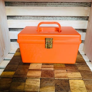 May include: An orange plastic toolbox with a handle and a brass-colored clasp. The toolbox is rectangular and sits on a wooden surface made of small, square tiles. The background is a wooden shelf.