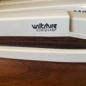 May include: Close-up of a white kitchen tool with the text "wiltshire STAYSHARP" in black. The tool has a dark brown wooden section and is set against a dark wooden surface.