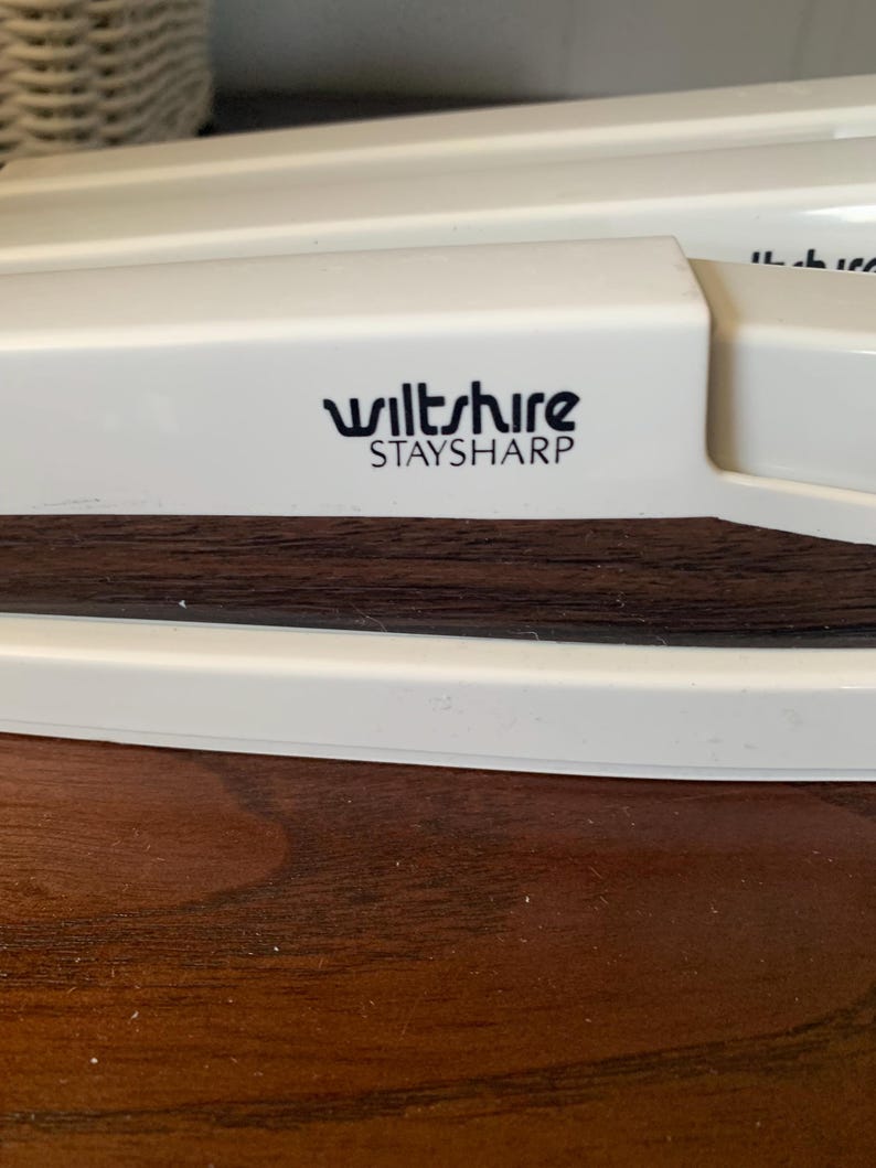 May include: Close-up of a white kitchen tool with the text "wiltshire STAYSHARP" in black. The tool has a dark brown wooden section and is set against a dark wooden surface.