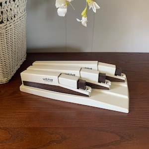 May include: A cream-colored Wiltshire StaySharp knife sharpener with three sharpening slots. The sharpener has a rectangular base and a brown handle. The Wiltshire logo is visible on the front. The sharpener is on a wooden surface.