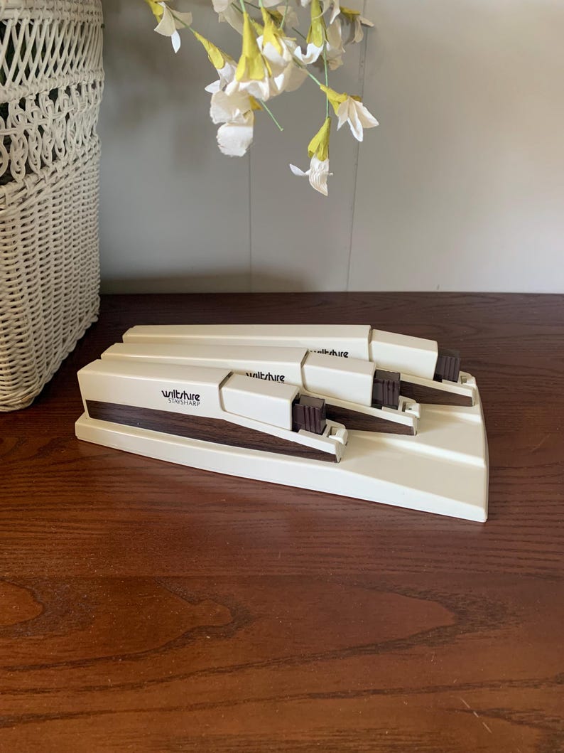 May include: A cream-colored Wiltshire StaySharp knife sharpener with three sharpening slots. The sharpener has a rectangular base and a brown handle. The Wiltshire logo is visible on the front. The sharpener is on a wooden surface.