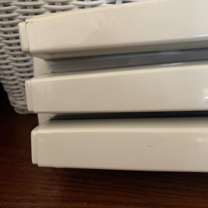 May include: Close-up of a stack of three off-white, rectangular objects with a glossy finish. The objects are stacked on top of each other, with a dark gray space between each. The objects are on a dark brown wooden surface.