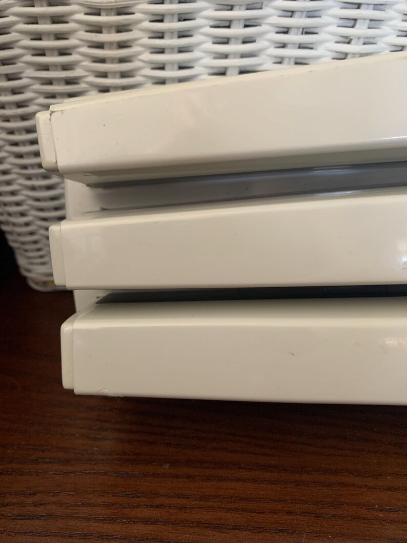 May include: Close-up of a stack of three off-white, rectangular objects with a glossy finish. The objects are stacked on top of each other, with a dark gray space between each. The objects are on a dark brown wooden surface.
