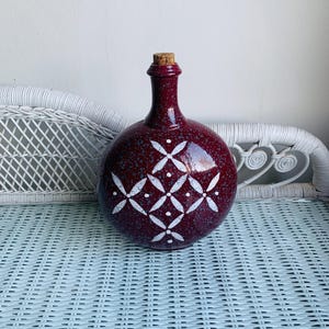 May include: A round, burgundy ceramic bottle with a cork stopper. The bottle features a white geometric pattern and a speckled finish. The bottle is likely used for decorative purposes or storing liquids.