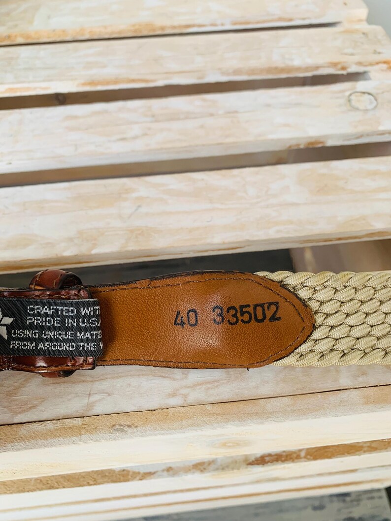 Puede incluir: Primer plano de un cintur&oacute;n tejido marr&oacute;n y beige. El cintur&oacute;n tiene una etiqueta de cuero con el n&uacute;mero "40 33502" en relieve. El cintur&oacute;n est&aacute; fabricado con orgullo en los EE. UU.
