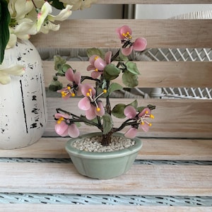 May include: A decorative bonsai tree sculpture featuring light pink translucent flower petals with yellow centers, green leaves, and dark brown branches. The tree is set in a small, light green ceramic pot filled with white gravel stones.