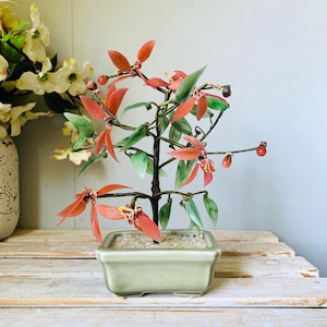 May include: A decorative bonsai tree with red and green glass leaves and flowers. The tree is planted in a square, light green ceramic pot filled with small white stones. The bonsai is displayed on a wooden surface.