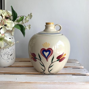 May include: A cream-colored ceramic jug with a handle and a lid. The front features a hand-painted design of a central blue and red heart topped with yellow rays, flanked by two stylized red and yellow flowers with green stems and leaves.