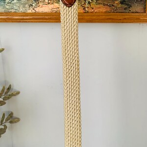 Puede incluir: Un cintur&oacute;n beige tejido con una punta de cuero marr&oacute;n. El cintur&oacute;n est&aacute; hecho de un material tejido apretado, y la punta de cuero tiene una apariencia texturizada. El cintur&oacute;n est&aacute; dise&ntilde;ado para usar con pantalones o faldas.