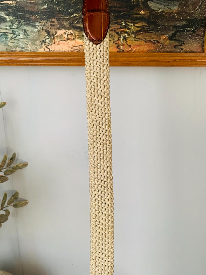 Puede incluir: Un cintur&oacute;n beige tejido con una punta de cuero marr&oacute;n. El cintur&oacute;n est&aacute; hecho de un material tejido apretado, y la punta de cuero tiene una apariencia texturizada. El cintur&oacute;n est&aacute; dise&ntilde;ado para usar con pantalones o faldas.