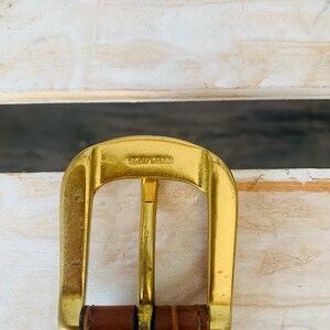 Puede incluir: Primer plano de una hebilla de cintur&oacute;n de lat&oacute;n macizo de color dorado con el texto "SOLID BRASS" estampado en la parte superior. La hebilla est&aacute; sujeta a un cintur&oacute;n de cuero marr&oacute;n. El fondo es una superficie de madera de color claro.
