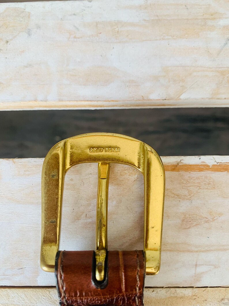 Puede incluir: Primer plano de una hebilla de cintur&oacute;n de lat&oacute;n macizo de color dorado con el texto "SOLID BRASS" estampado en la parte superior. La hebilla est&aacute; sujeta a un cintur&oacute;n de cuero marr&oacute;n. El fondo es una superficie de madera de color claro.