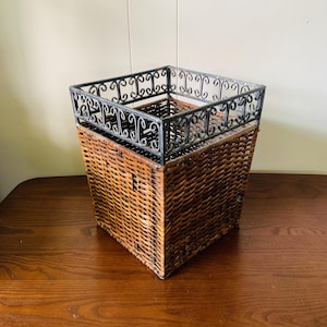 May include: A square wicker basket with a decorative black metal top. The basket is made of woven brown wicker and has a dark metal frame with an ornate design. The basket sits on a wooden surface.