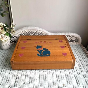 May include: A rectangular wooden box with a hinged lid featuring a painted dark blue cat silhouette in the center. The box is decorated with a small pink bow above the cat and six pink hearts with white dotted outlines surrounding the cat.