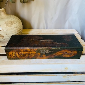 May include: A rectangular wooden box with a dark, ornate lid featuring gold and brown swirling designs. The box is on a white wooden crate, with a cream-colored vase and decorative branches in the background.