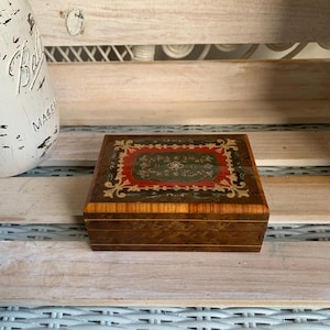 May include: A rectangular wooden box with a polished finish featuring a dark brown wood grain. The lid displays a decorative inlay with a central dark green floral motif surrounded by a red border and intricate cream-coloured scrollwork patterns.