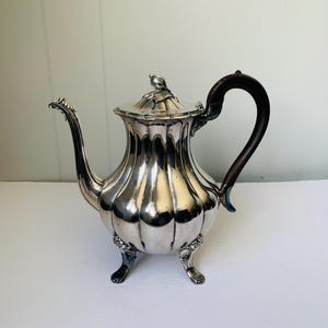 May include: A silver teapot with a dark brown handle and three feet. The teapot has a fluted design and a decorative knob on the lid.