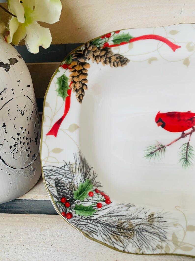 May include: White ceramic serving dish with a gold rim. The dish features a festive Christmas design with red cardinals, pine branches, holly berries, pine cones, and red ribbon.
