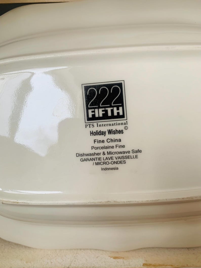 May include: White ceramic serving dish with a black and white logo that reads "222 FIFTH PTS International Holiday Wishes Fine China Porcelaine Fine Dishwasher & Microwave Safe GARANTIE LAVE VAISSELLE / MICRO-ONDES Indonesia"
